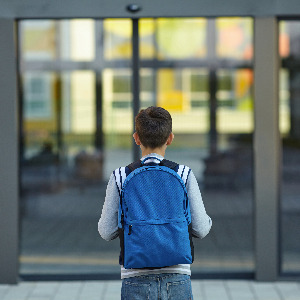 An elementary school student stands before school doors about to enter. He has a blue backpack.