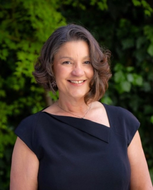 Portrait of Marlene Sallo, a Latino woman with short dark hair. She is wearing a black top and stands outside in front of green leaves and foliage.