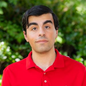 Eden Alon. He is a young, Middle Eastern man with olive skin, dark hair, and brown eyes. He has a slight smile on his face and a curious facial expression. He is wearing a red polo shirt. The background is blurry but appears to consist of green bushes or trees.