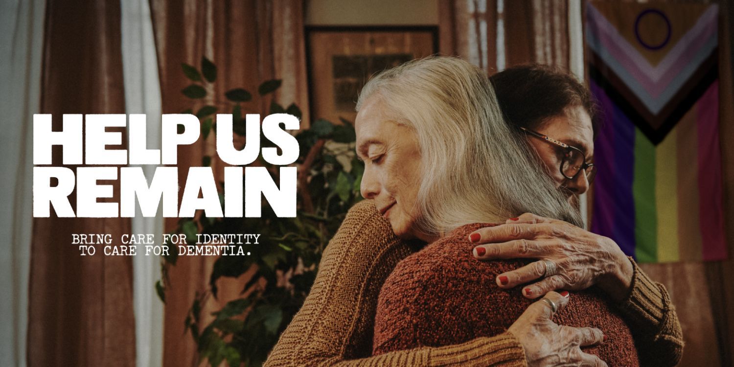 Help us Remain. Bring care for identity to care for dementia. Two older adults hug. Behind them hangs the Intersex-Inclusive Progress Pride flag.