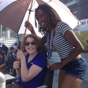 Image of the author, much younger. They are white with shoulder-length brown hair. They are sitting in a wheelchair and holding an umbrella. Standing next to them is Tamita, a Black woman with shoulder-length locs. She is smiling and wearing a T-shirt and shorts. There is a gold medal around her neck. Just out of the frame is Tamita’s prosthetic leg on her right side. Tamita had just changed out of her running blade into her regular prosthetic leg.