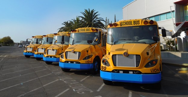 Five electric buses are parked in a lot in front of a school.