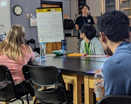 This image shows a classroom setting with a diverse group of students seated at tables, attentively facing a speaker at the front of the room. The speaker is standing near a flip chart with several topics listed, such as ‘Asking Questions’, ‘Expressing Concerns’, ‘Asserting Preferences’, ‘Active Listening’, ‘Building Support Networks’, and ‘Not Self-Advocacy’. The classroom has various educational materials, including a periodic table of elements, cabinets with glassware, and a clock on the wall. The atmosphere appears to be inclusive and focused on learning about communication and advocacy skills