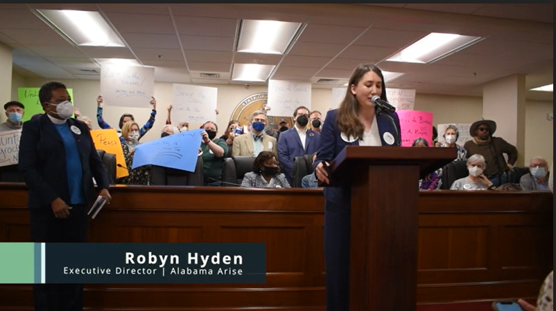 A room shows a group of people holding colorful signs in the background. In front a woman speaks at a podium and another stands to the side.