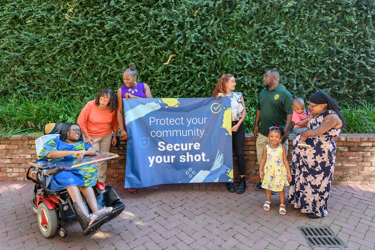 Diverse group of disabled people pose with a banner that reads, 'protect your community, secure your shot!'