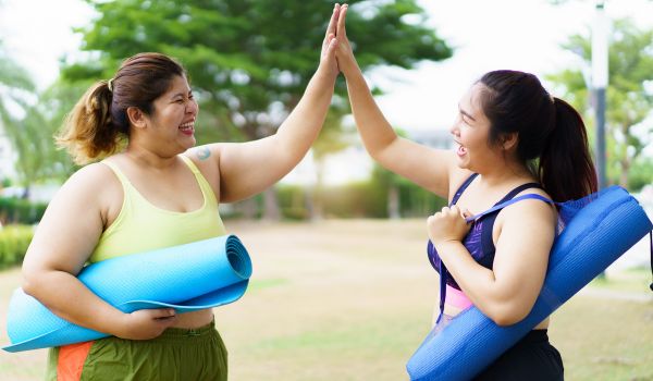Two young adult women high five together in a park, with their yoga mats on their shoulders