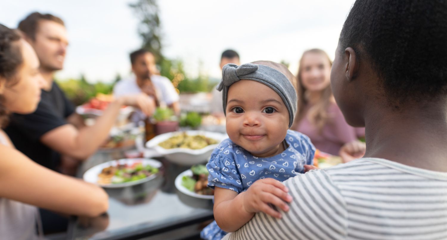 A baby smiles at the camera while being held at an outdoor meal with family and friends