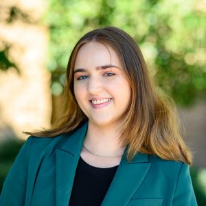 Isabelle, a young woman with straight light brown hair is shown smiling in an outdoor setting. She is wearing a dark green blazer over a black top. The background features soft-focus greenery and sunlit trees. Her posture is relaxed, and she appears confident and approachable.