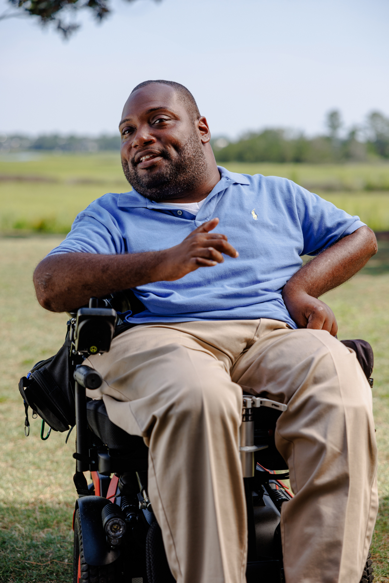 Alex, a Black man in a power wheelchair speaking while outside with a marsh behind him.