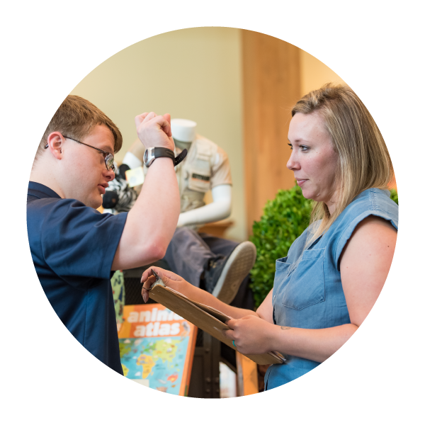 Young man with a developmental disability stands to the left talking to a woman with a clipboard to the right. They are in a store setting where the young man works.