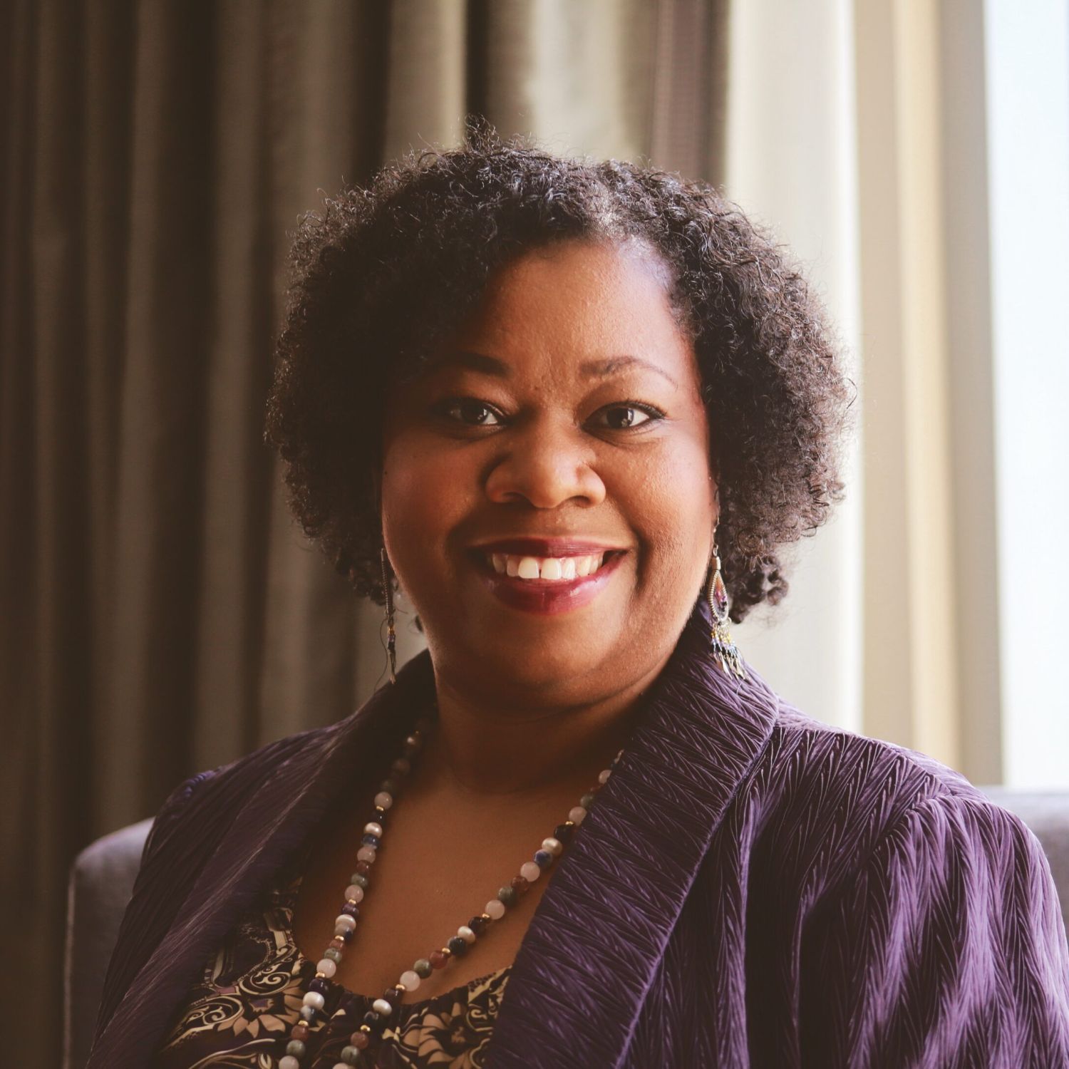 A Black woman wearing a beaded necklace and purple cardigan smiles at the camera