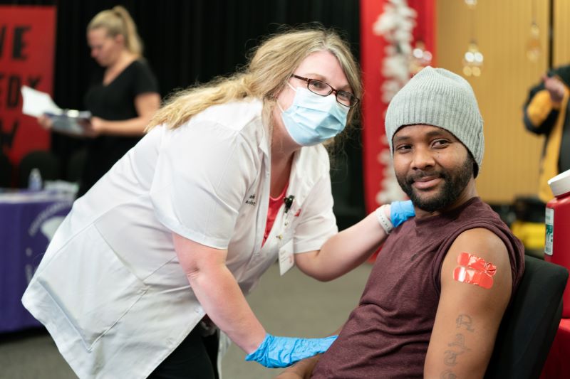 A nurse poses with a patient in a chair- a white woman in a white lab coat and a black man with bandaids on his arm.