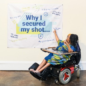 Ebony- a young Black woman in a power wheelchair, points to a sign that reads, 'Why I secured my shot.'