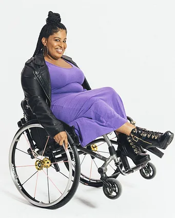 Tatiana Lee, a black woman wearing a purple dress and high heel combat boots doing a wheelie with her wheelchair while smiling.