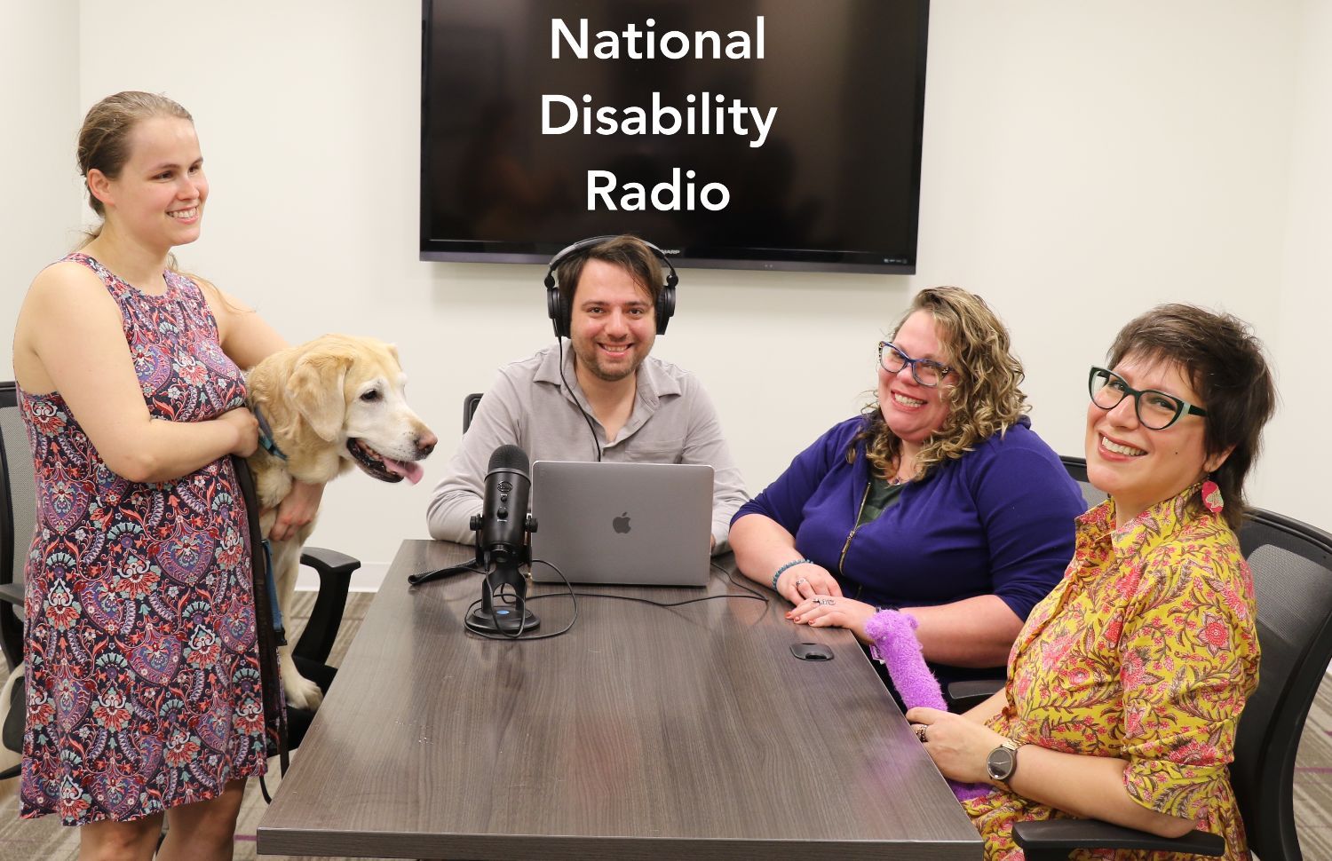 [3:07 PM] Jack RosenThe hosts of the podcast from left to right: Stephanie, Jack, Michelle and Raquel, gather around a conference table. Stephanie is standing while holding her guide dog Nala who has a mic in front of her. Above Jack's head on a TV are the words 