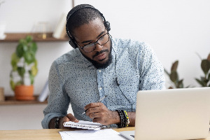 A black man sitting in front of a laptop wearing headphones and taking notes.