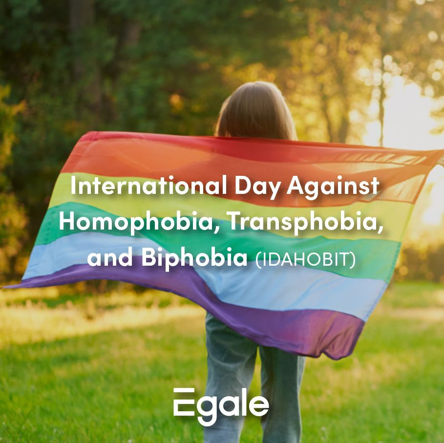 Person holding a rainbow pride flag behind their back. In front of the flag white text reads International Day Against Homophobia, Transphobia, and Biphobia (IDAHOBIT)