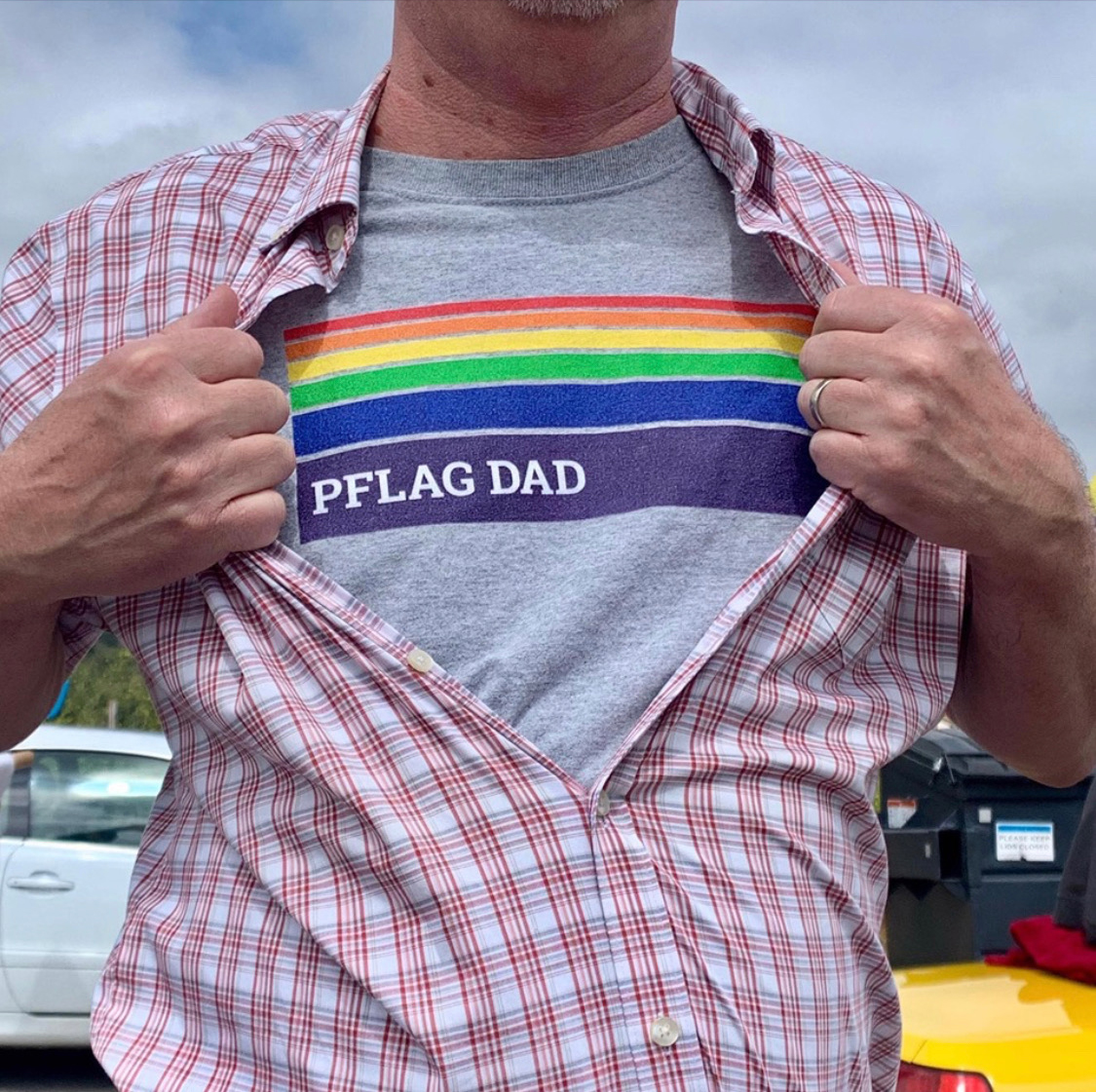 A male-presenting person opening their button-down shirt, revealing a Pride image that says, 
