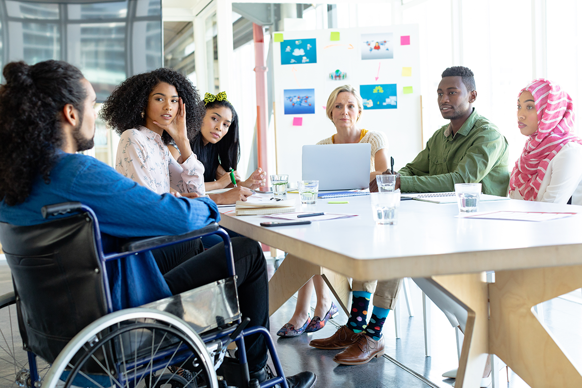 Group of people sitting in a meeting room looking at a wheelchair-using person speak.