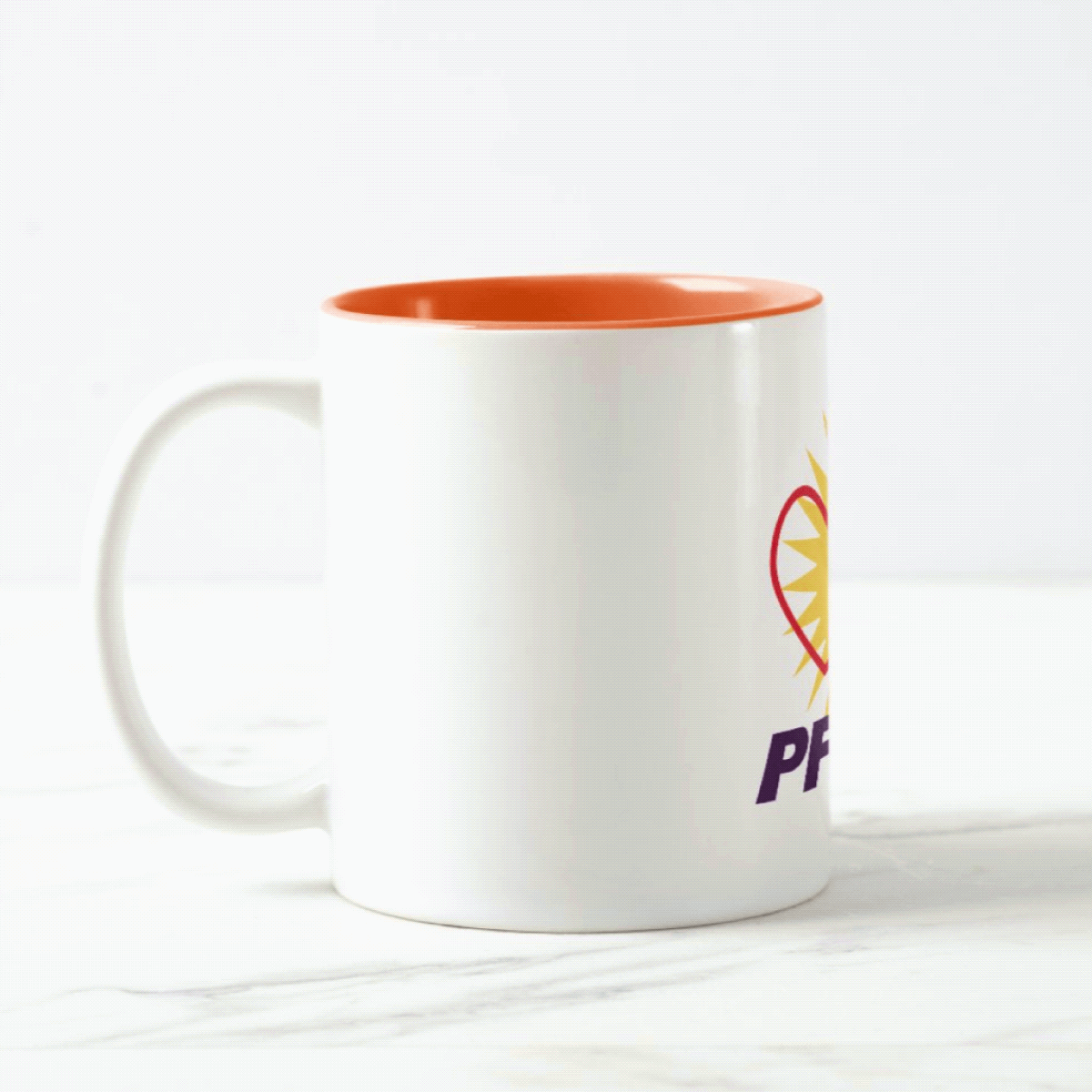 A gif of a PFLAG National ceramic mug on a white surface rotating back and forth. The mug is white on the outside and orange on the inside.