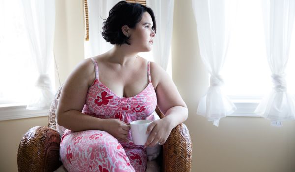 A woman holds a coffee mug while sitting in a chair and looking out a sunny window.