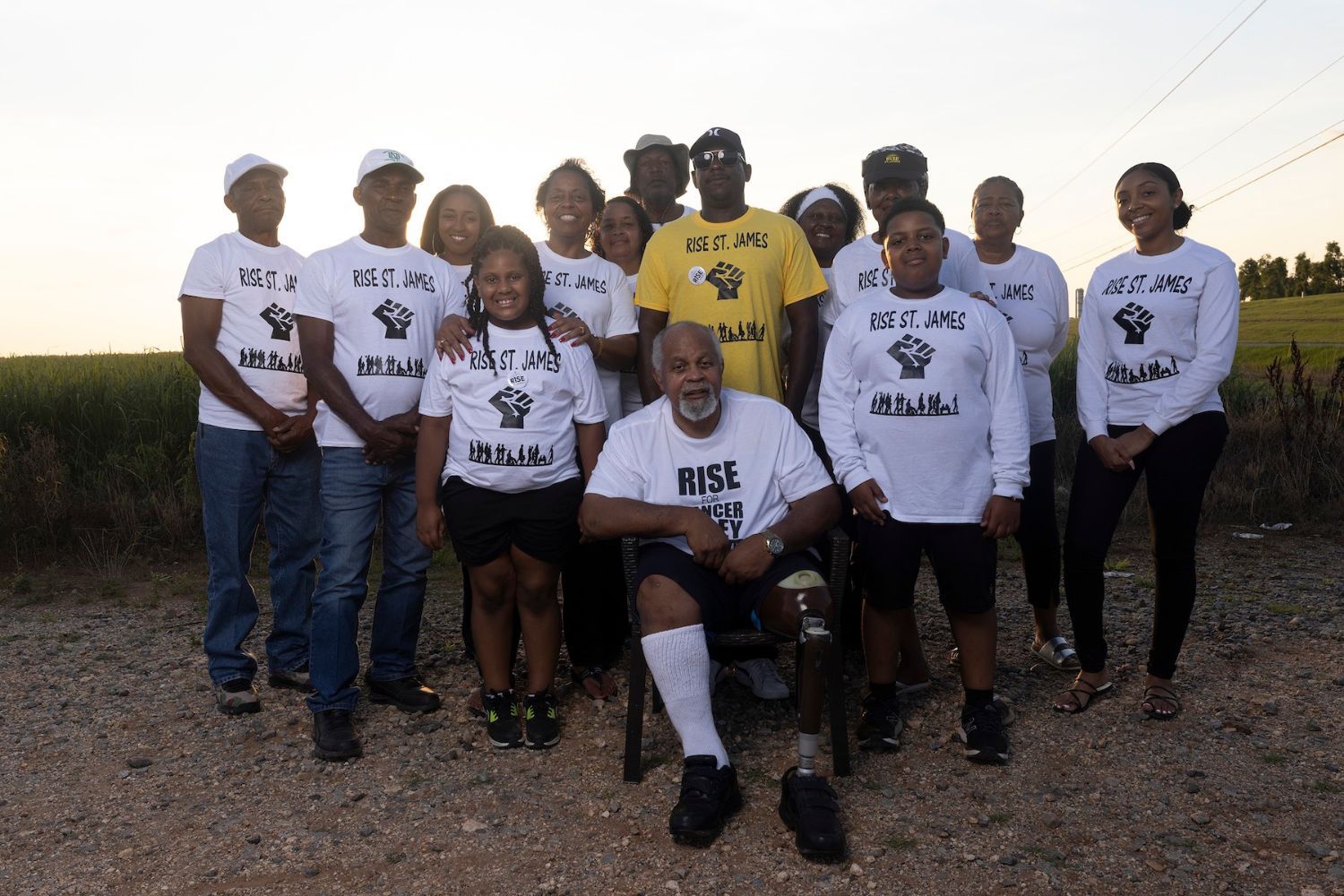 A group of people standing in a field lit by sun behind them. Everyone is wearing t-shirts saying Rise St. James with a raised fist in the center of the shirt.