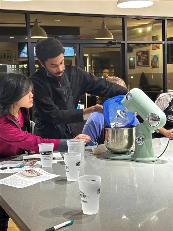 A group of people in a kitchen, learning how to bake or cook. A man in a black jacket is pouring something into a big mixing bowl that is attached to a light green stand mixer. A woman in a red shirt is watching closely. There are papers, cups of water, and other baking tools on the table. The room looks bright and clean