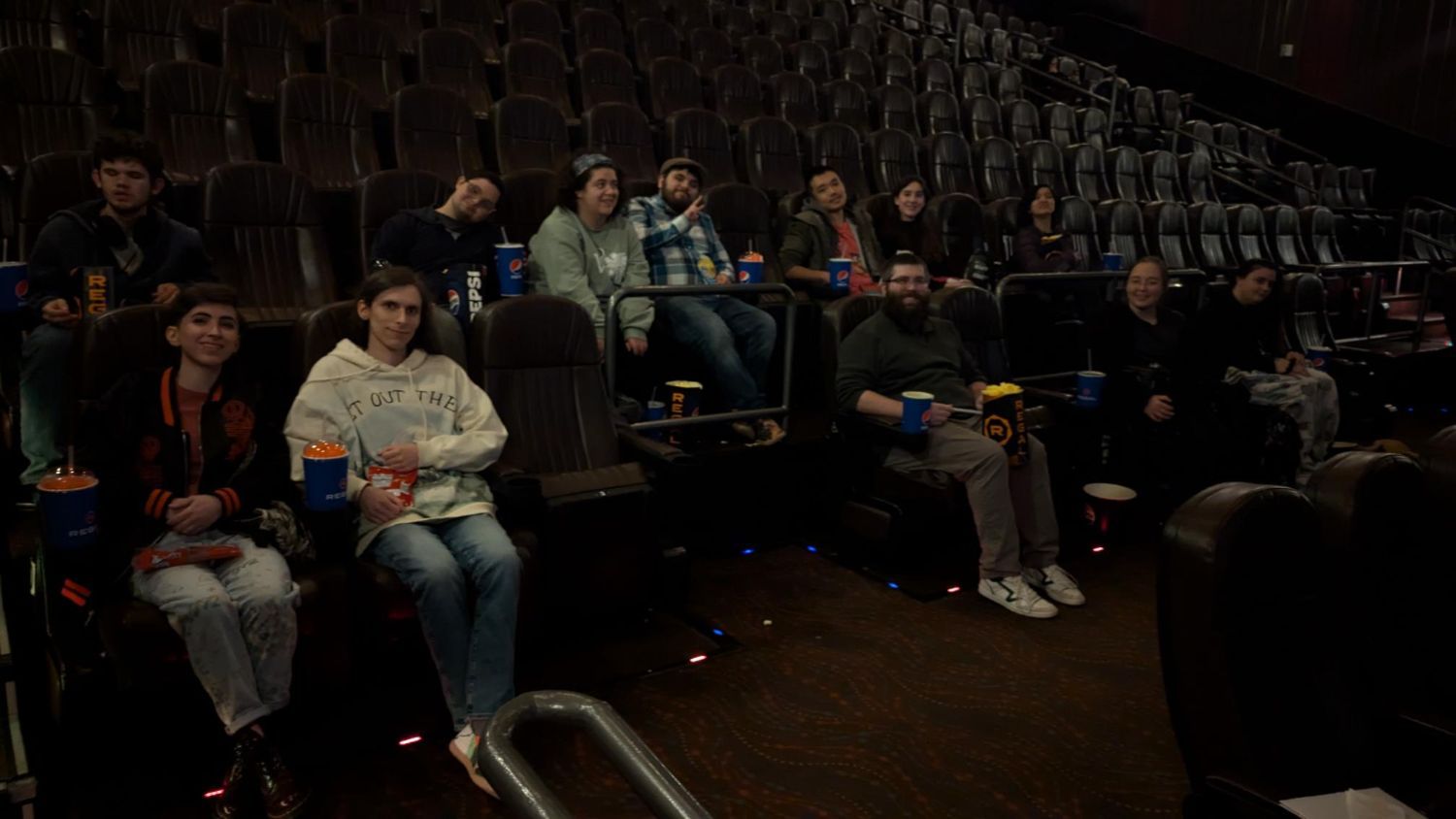 A small group of people sitting in a movie theater. There are a lot of empty seats around them. They look happy and relaxed, and some are smiling at the camera. Many of them are holding snacks like popcorn, drinks, and candy. Some people are leaning back in their seats, and others are sitting up. The theater is a little dark, like it usually is before a movie starts.