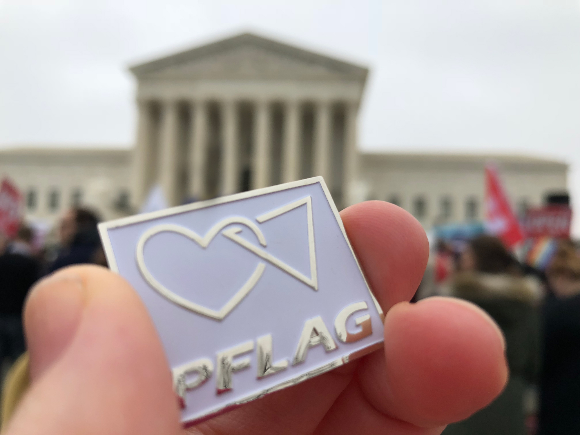 The Supreme Court building in the background with a hand holding a PFLAG pin in the foreground.