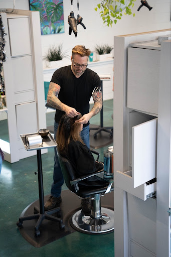 A white man is standing up, wearing a black t-shirt. He styles the hair of a woman with dark hair who sits in a chair.