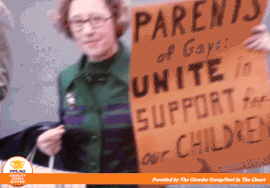 A video loop of Jeanne Manford, a white woman with brown hair in a green dress, marching holding a sign on brown posterboard with the words "Parents of Gays Unite In Support For Our Children" in black lettering.