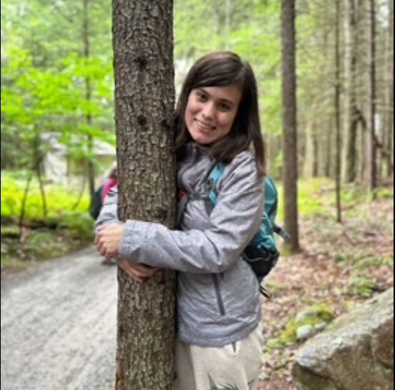 Avery Grace Arthur, a young white woman with long hair, stands smiling and hugging the trunk of a tree. She is wearing a grey jacket, blue backpack, and white pants. There are more trees and some grass in the background. To her left, there is a gravel road.