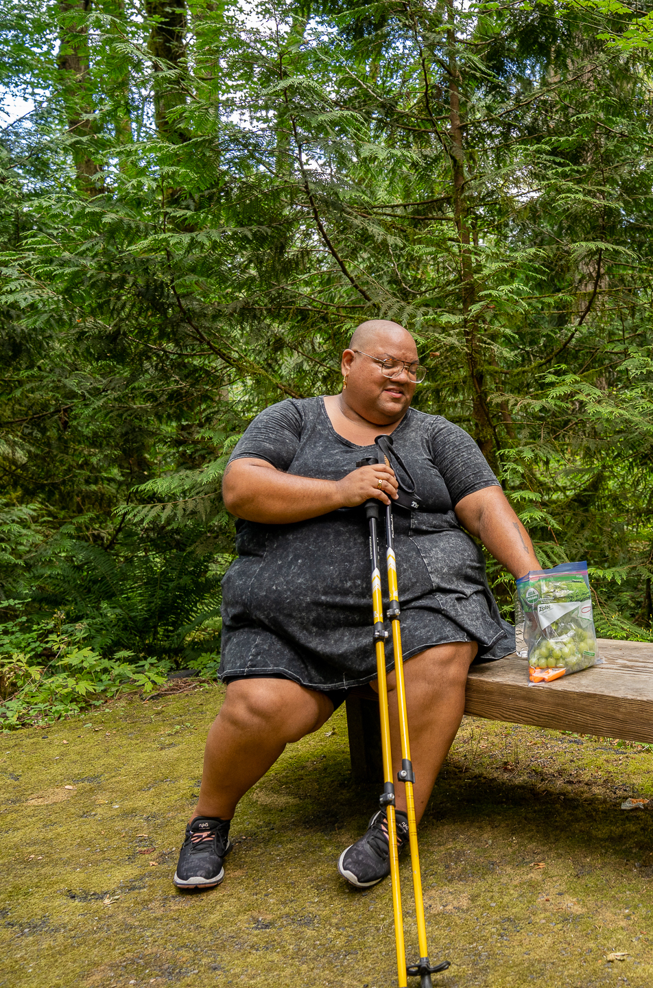A disabled nonbinary Black woman sits on the side of a hiking trail eating grapes.