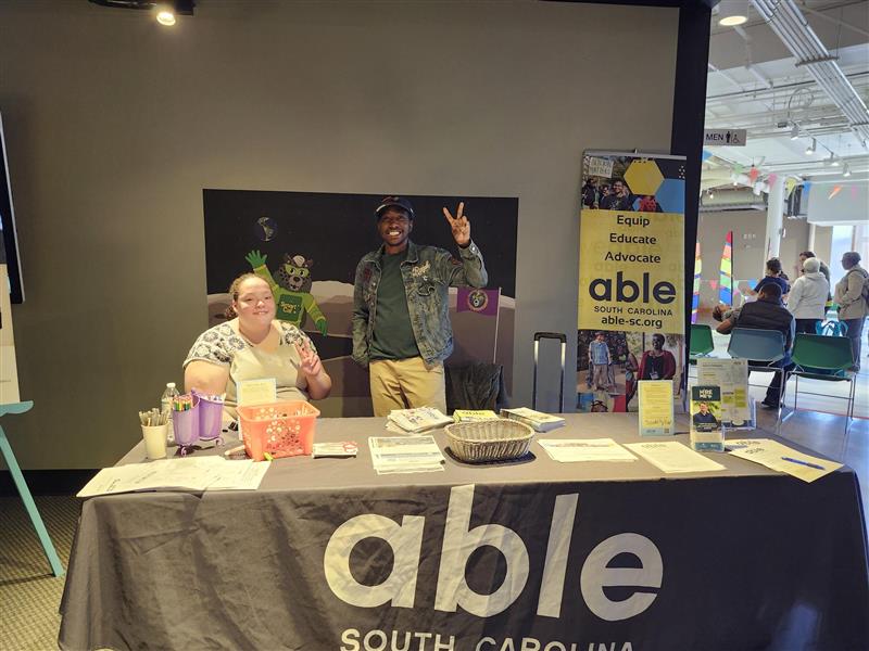 Youth Transition Specialist, Donovann Sumter, and AmeriCorps Member, Dajha Pridgen, smile at Disability Celebration Day at the museum, next to an Able SC table and banner.