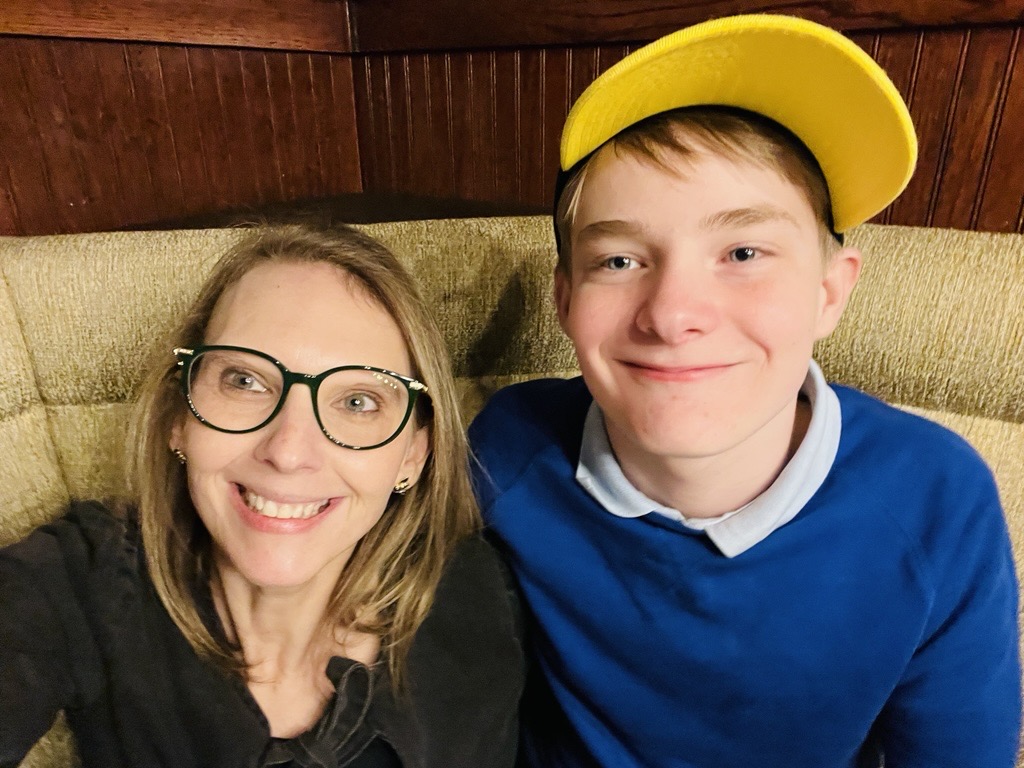 Kimberly and her son smile together. Kimberly is a white woman with glasses. Carter is a teen boy wearing a yellow hat.