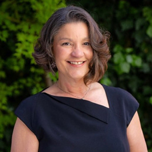 Portrait of Marlene Sallo, a Latino woman with short dark hair. She is wearing a black top and stands outside in front of green leaves and foliage.