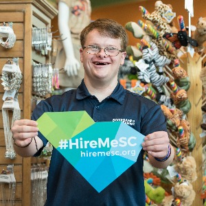 A white man with Down Syndrome working in a shop holds a sign that reads, 'Hire Me SC.'