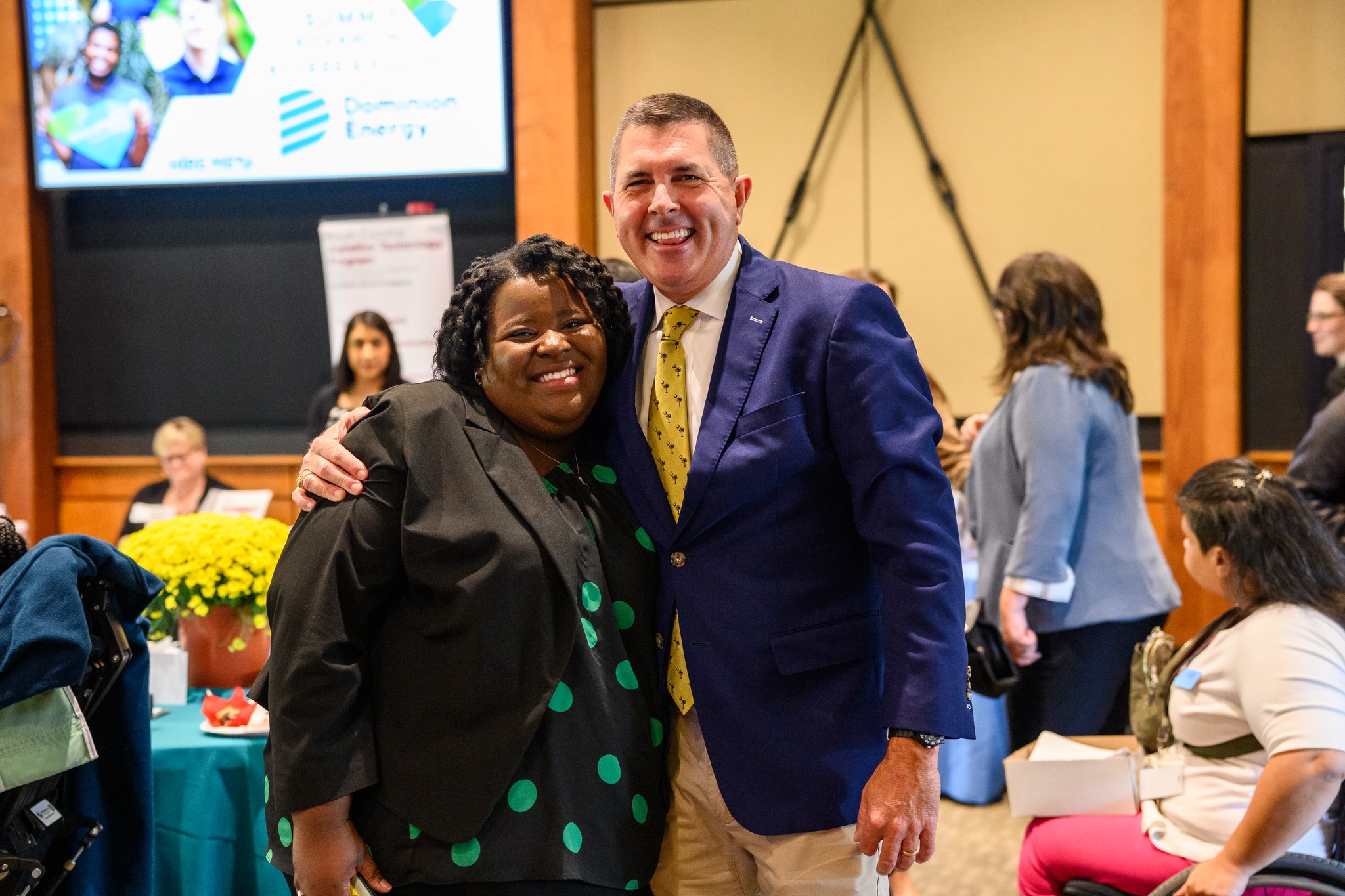 Event Keynote speaker, Keller Kissam, Dominion Energy President. He is a white man wearing a blazer. He stands side hugging an event participant, a Black woman wearing a black blazer. They both are smiling.