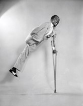 Black and white image of Heard, a black man with one leg and one arm. He is wearing a suit and tap shoes. He is smiling and jumping in the air, with a crutch held in his left arm allowing him to hold his position mid-air.